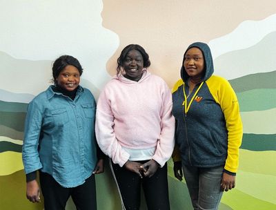 Three women stand in front of a wall painted with mountain landscape art.