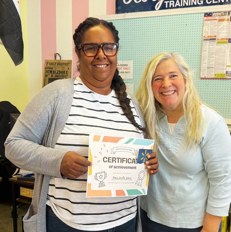 Two women smile, one holding a certificate. They stand inside a training center with pink and blue walls.