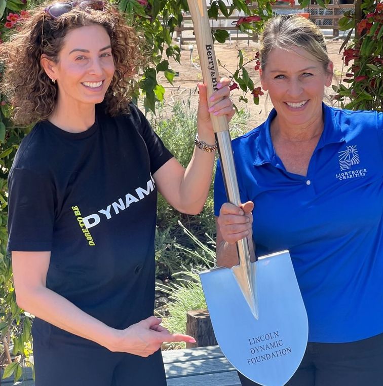 Two women holding a shovel, smiling outdoors. One points, blue shovel has logo, and is inscribed 