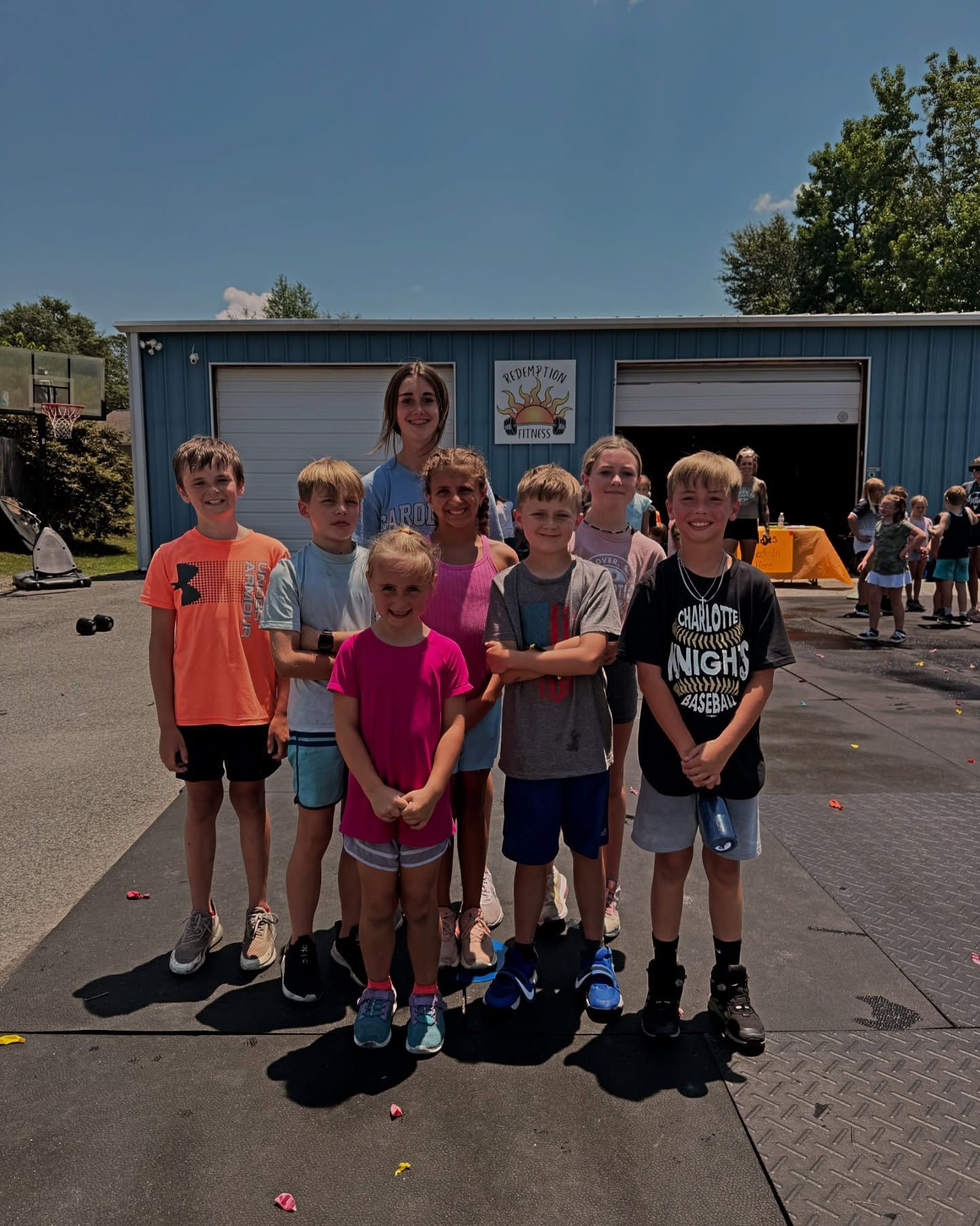 Group of children posing outdoors in front of a blue building on a sunny day.