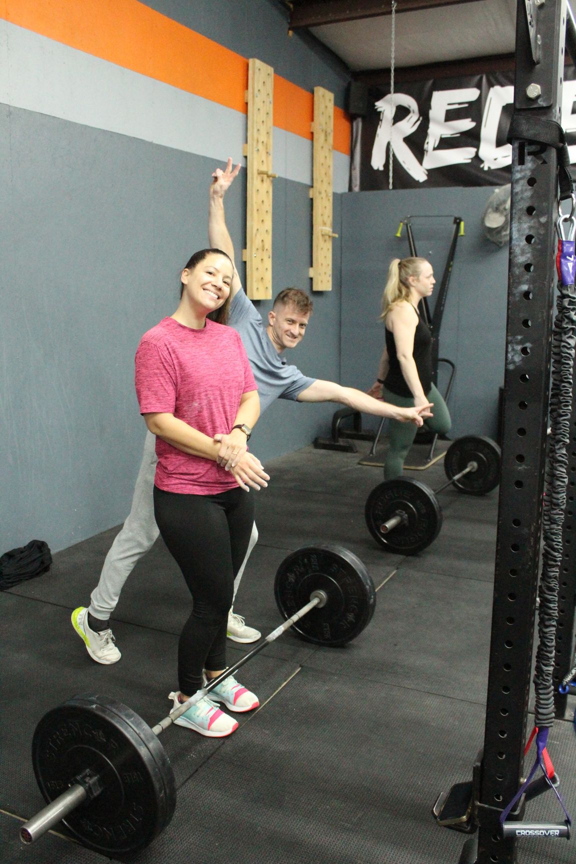 People smiling during a group workout in a gym with barbells and a REI sign on the wall