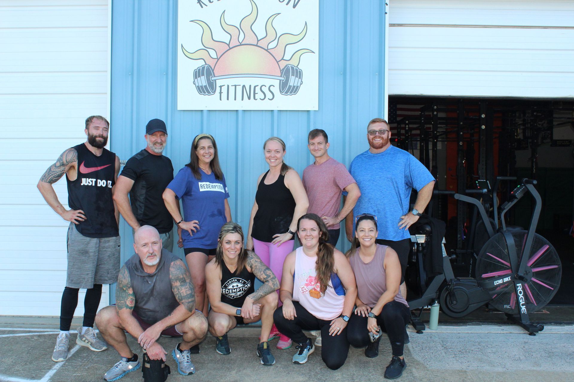 Group of people posing outside a fitness gym under a sun logo, with bikes and a motorcycle nearby