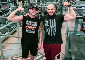 Two men flexing and smiling in a gym, standing beside workout machines.