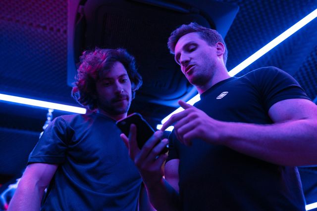 Two men looking at a phone under blue-purple neon lights in a dark indoor setting