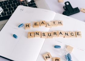 Scrabble tiles spelling “health insurance” on a desk with blue pills and papers