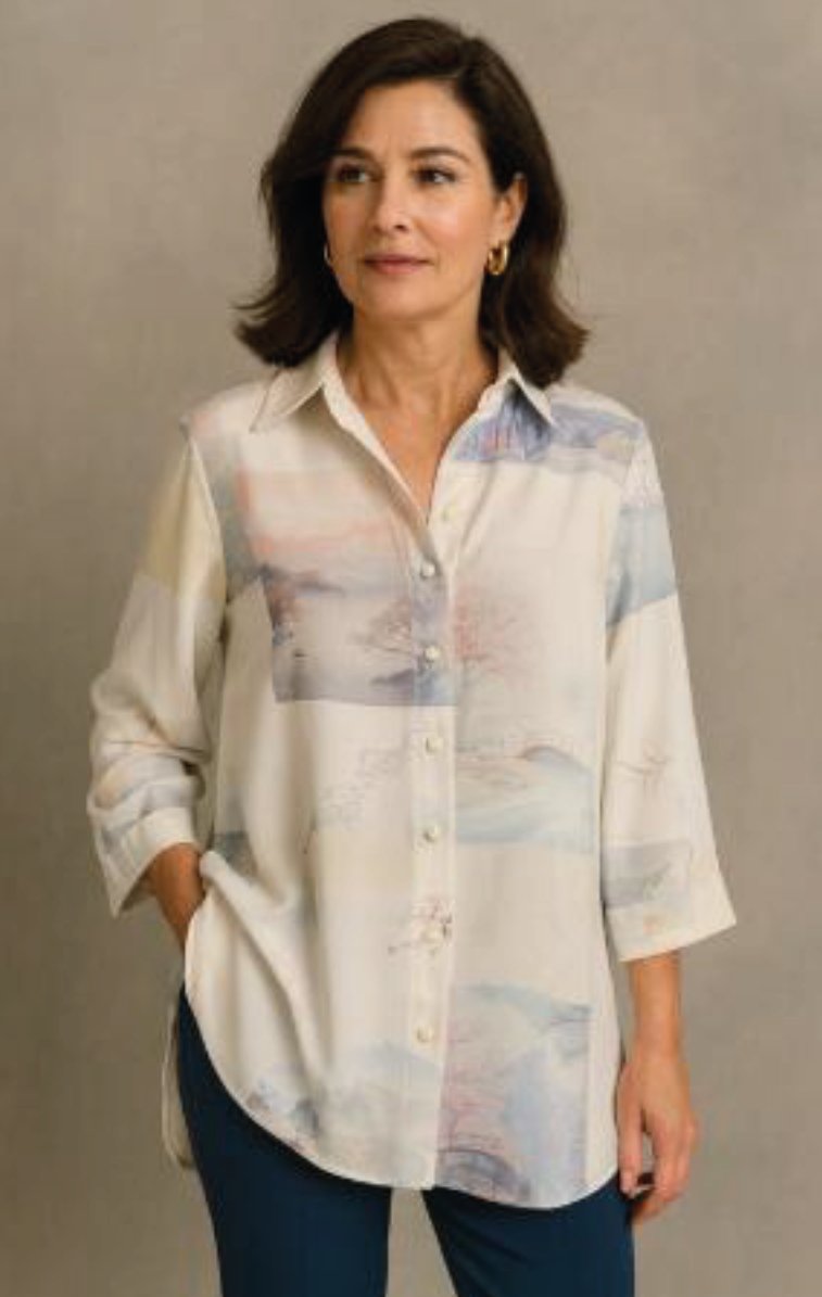 Mujer de pie frente a un fondo beige, vestida con una blusa blanca estampada en tonos pastel y vaqueros oscuros.