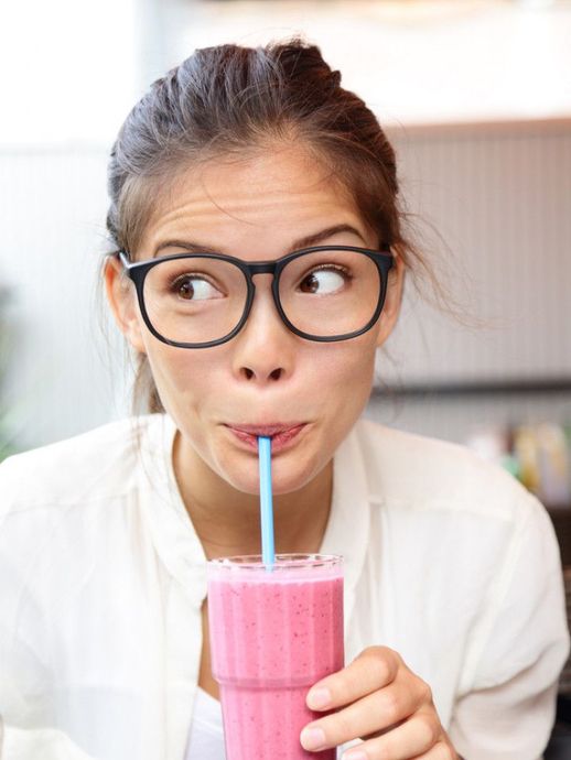 Woman with glasses sipping a pink smoothie and glancing sideways in a bright kitchen
