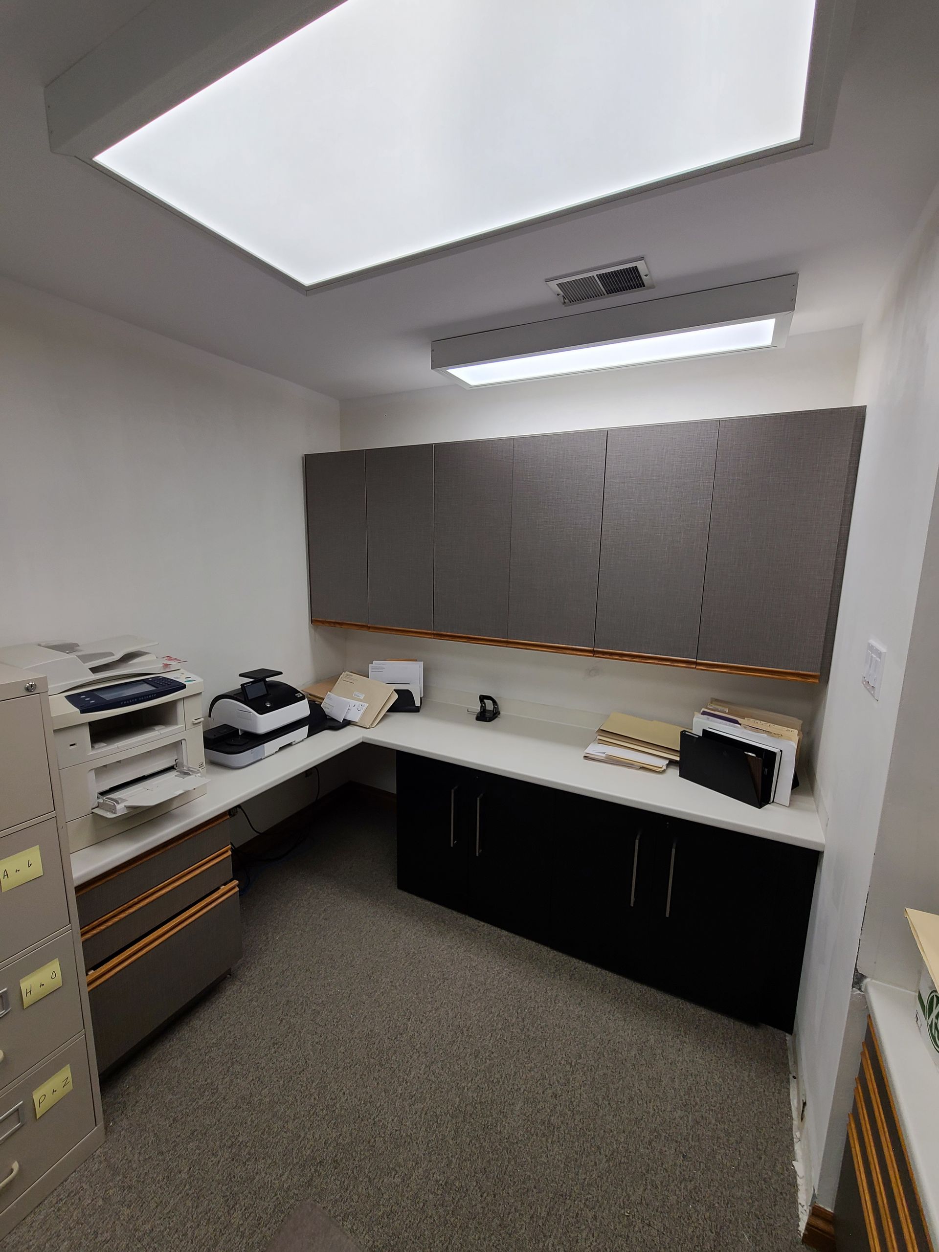 wrapped Office space with built-in desk, cabinets, and overhead lighting; filing cabinet on left.