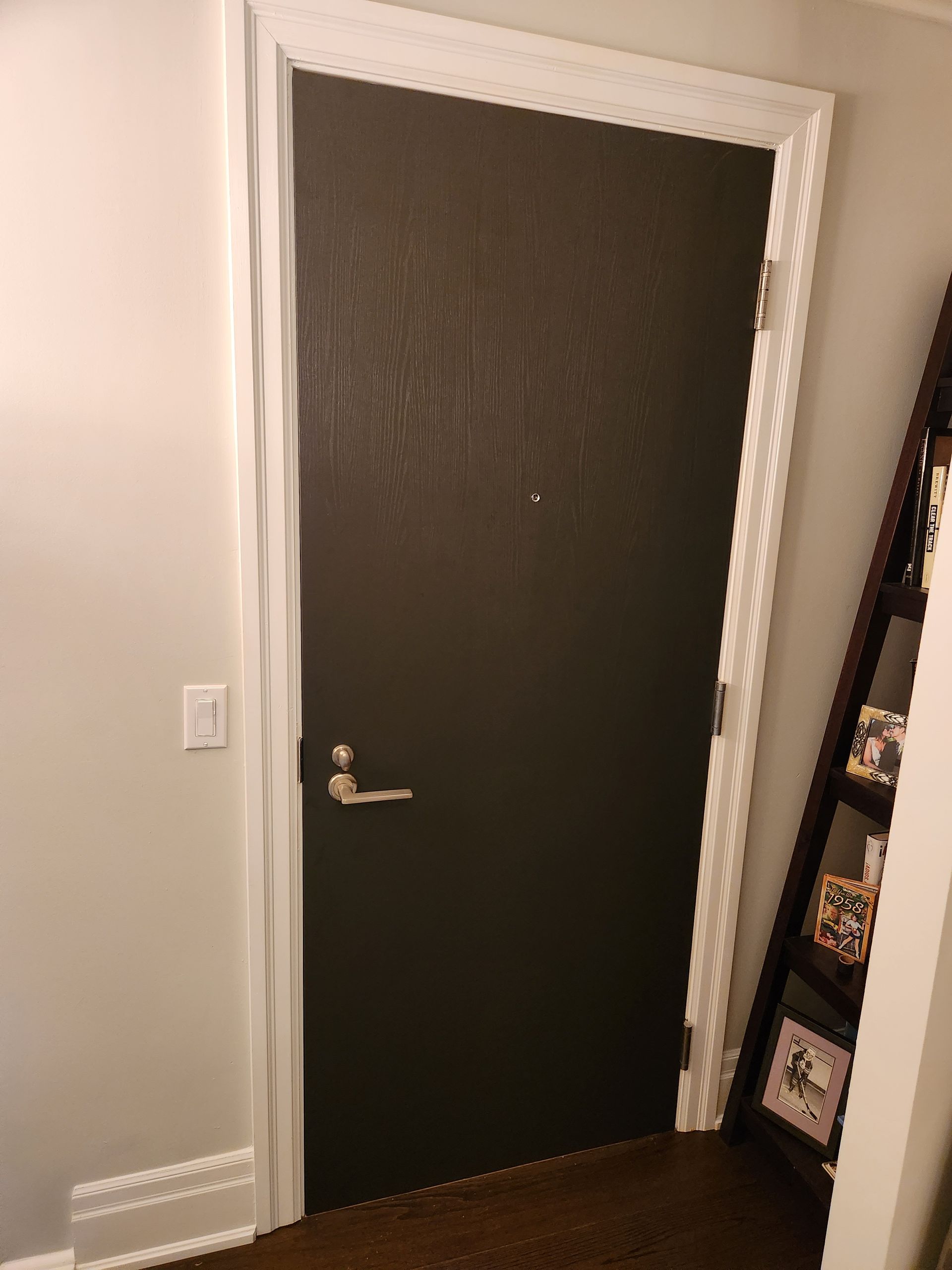 Dark brown door with white trim, silver handle, and peep hole. Leaning bookshelf to the right.