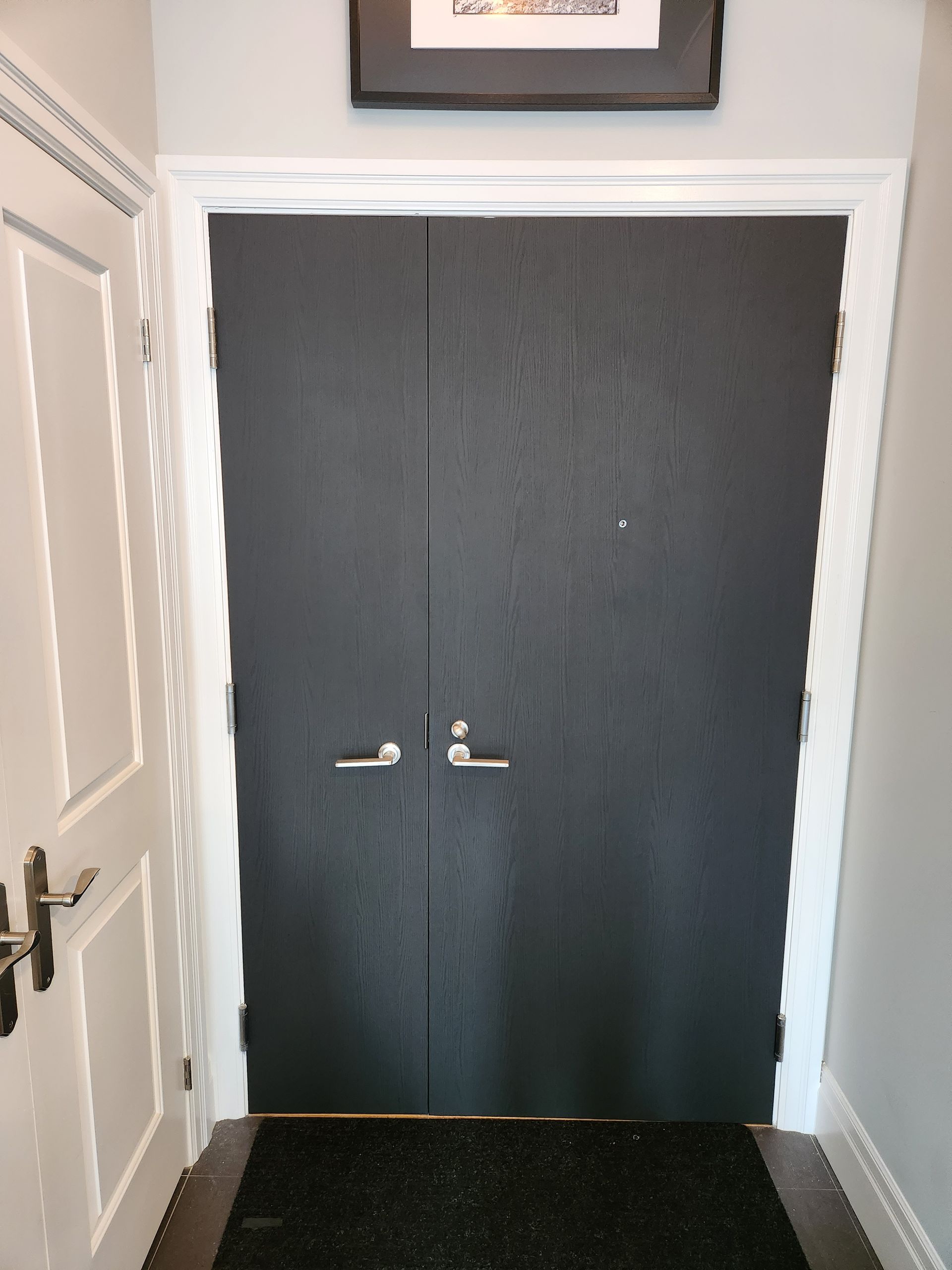 Black door with silver handles, doormat, white trim and frame, under a framed picture.