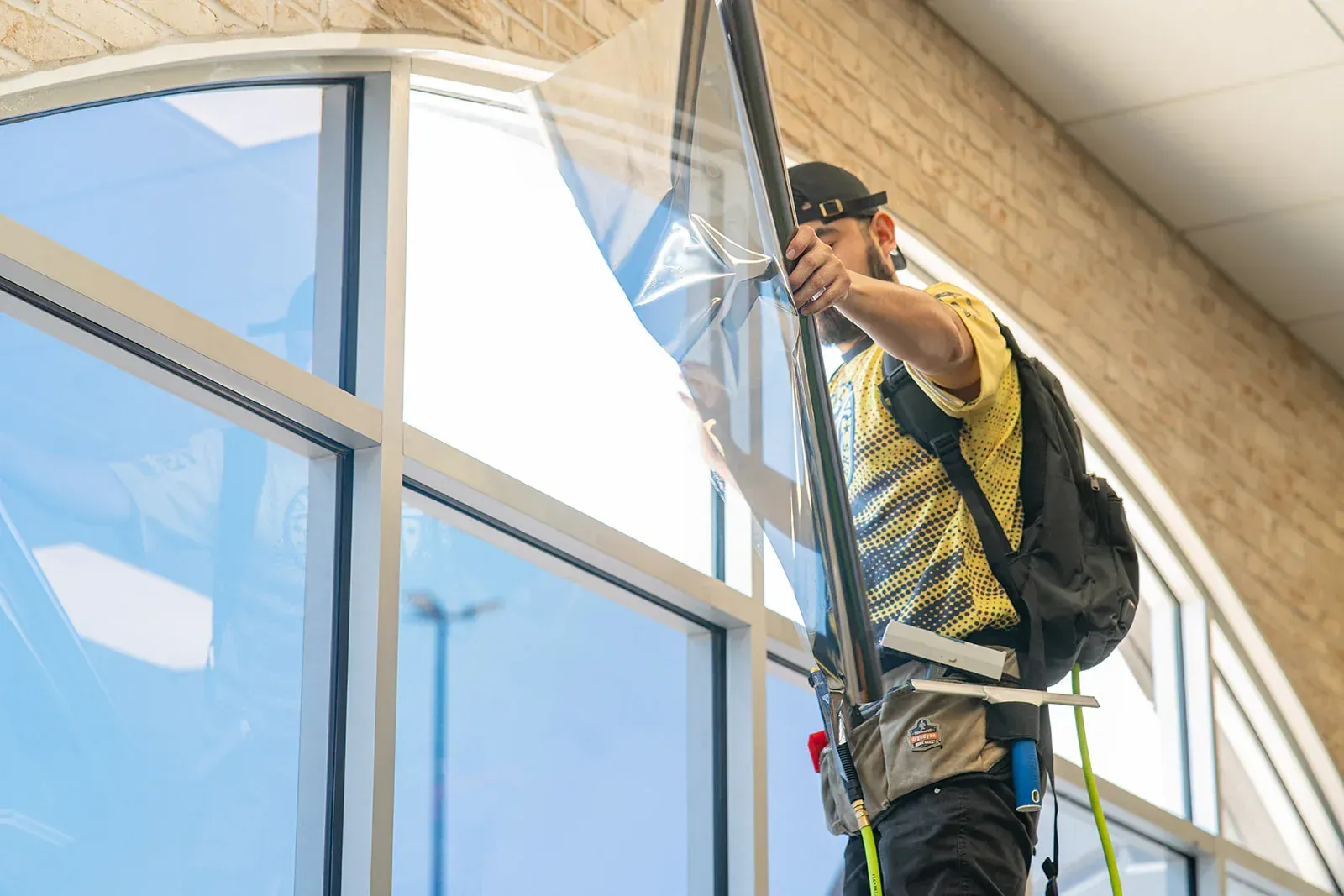 Man installing window film on a large arched window, wearing safety harness and backpack.