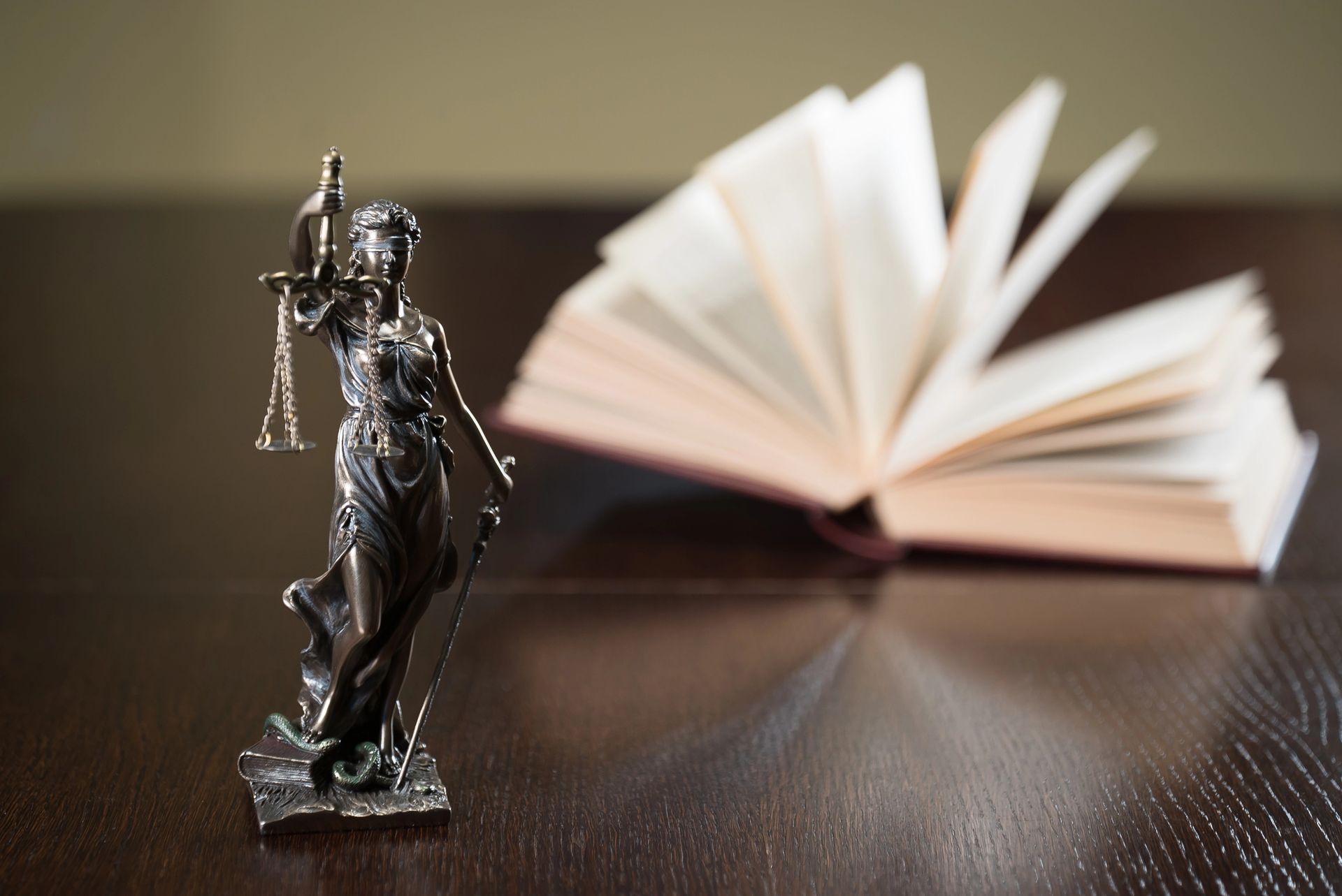 Una statua metallica della dea della giustizia che regge una bilancia si erge su un tavolo di legno scuro accanto a un libro aperto.