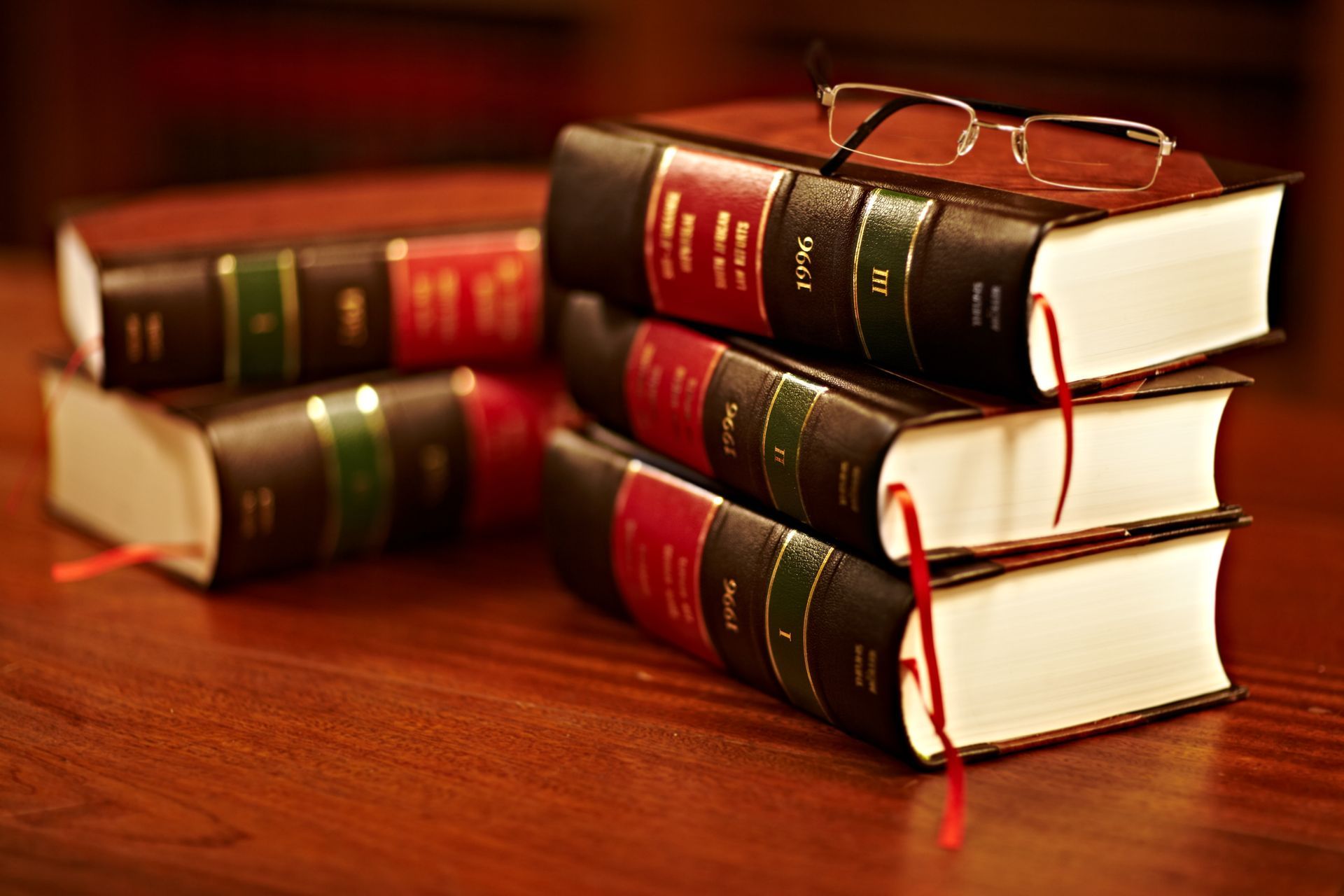 Una pila di tre voluminosi libri rilegati in pelle su una superficie di legno, con un paio di occhiali da lettura appoggiati sul libro in cima.