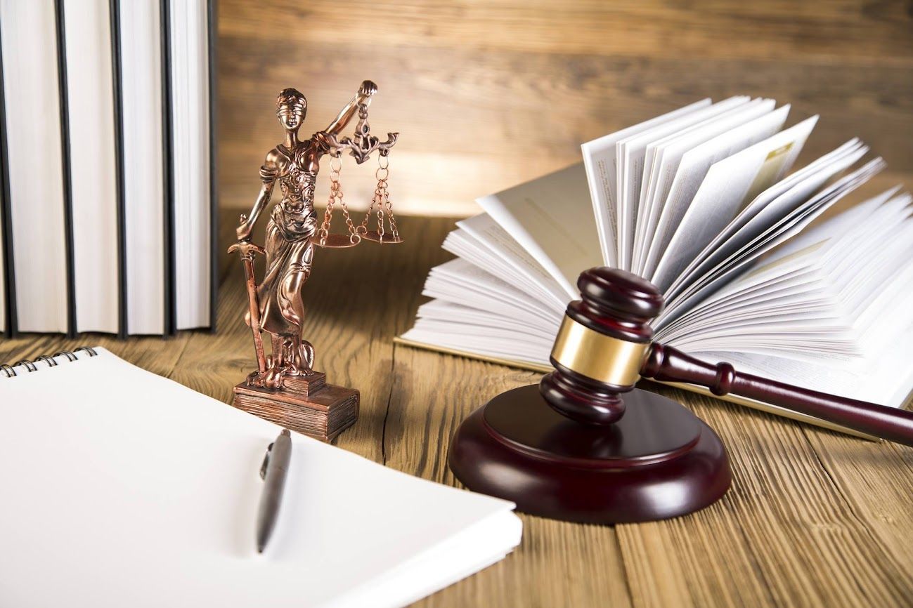 Una statua della dea della giustizia, un martelletto di legno, un libro aperto e un blocco note con una penna su una scrivania di legno.