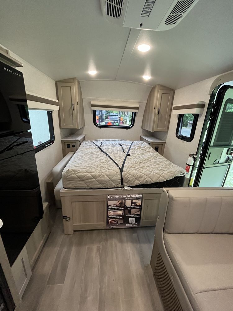 Inside a modern RV bedroom with a large bed, beige cabinets, and windows along the walls.
