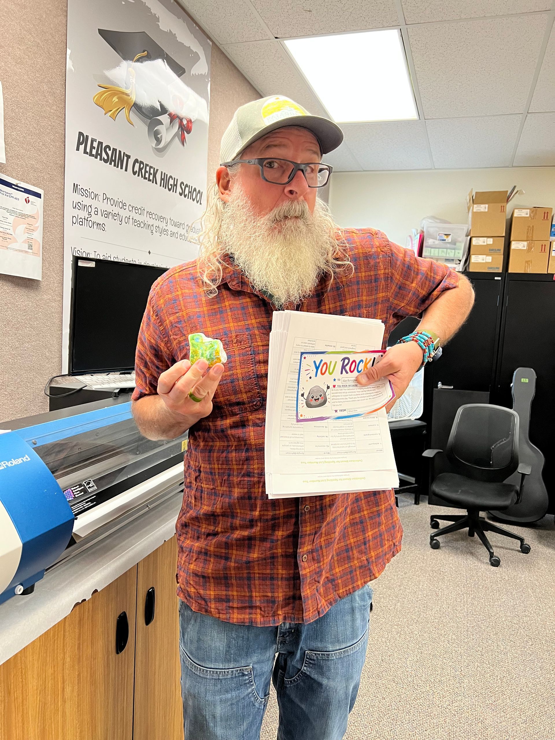 Man in plaid shirt holding a small toy and a booklet beside a large printer in an office workshop