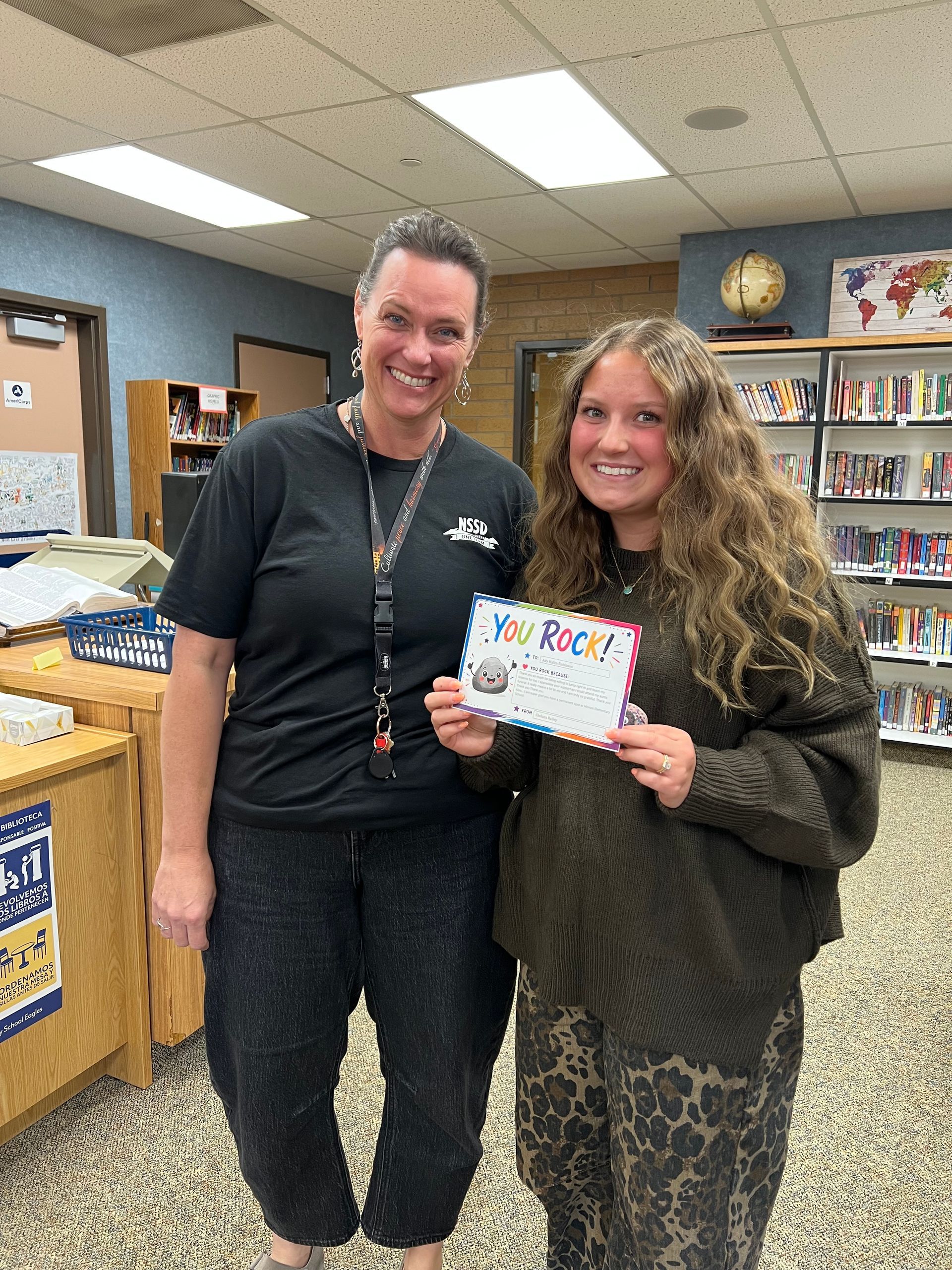 Two smiling people in a classroom hold a colorful sign, standing near bookshelves and a whiteboard.
