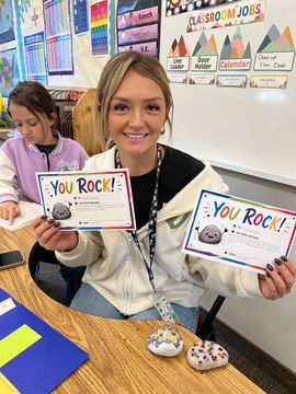 Student in a classroom holding two “You Rock!” certificates and smiling at the camera.