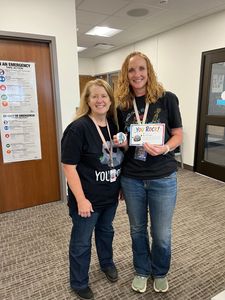 Two people in black shirts smiling in a hallway, holding a small sign and gift items.