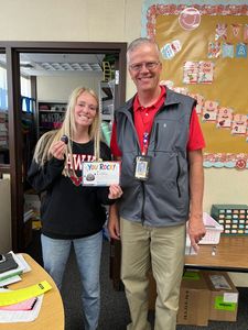 Two people smiling in a classroom, holding a colorful card and standing beside a bulletin board.