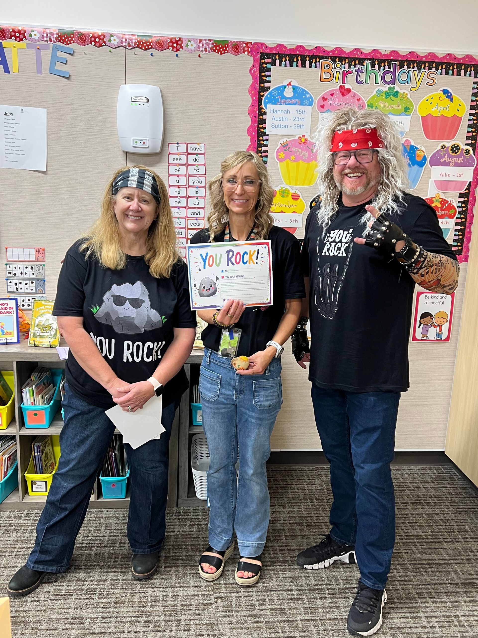 Three people smiling and holding a certificate in a classroom decorated with colorful posters.