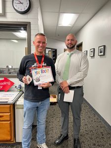 Two men in an office hallway; one holds a colorful award certificate.
