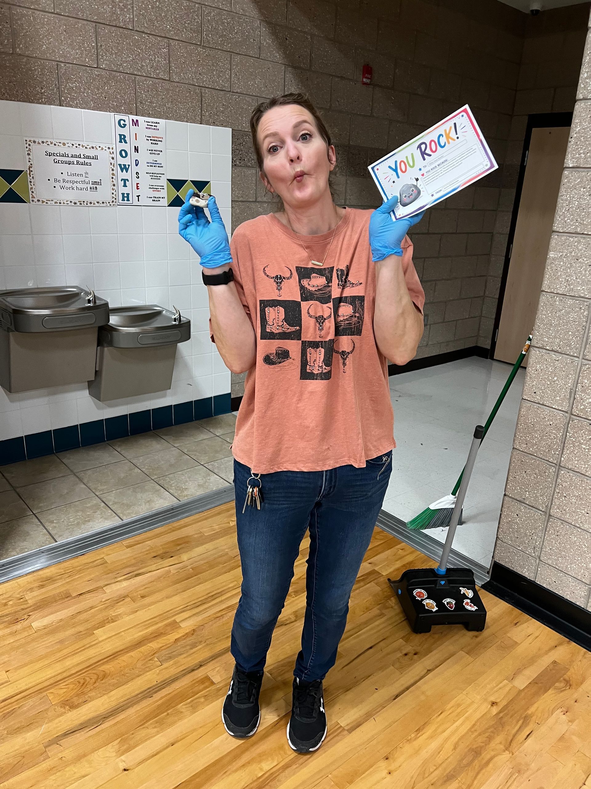 Woman in a gym holding a colorful sign and a blue item, wearing gloves and standing on a wood floor.
