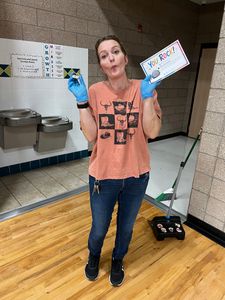 Woman in a hallway holds up a small sign and a blue object, wearing blue gloves and standing on a gym floor.