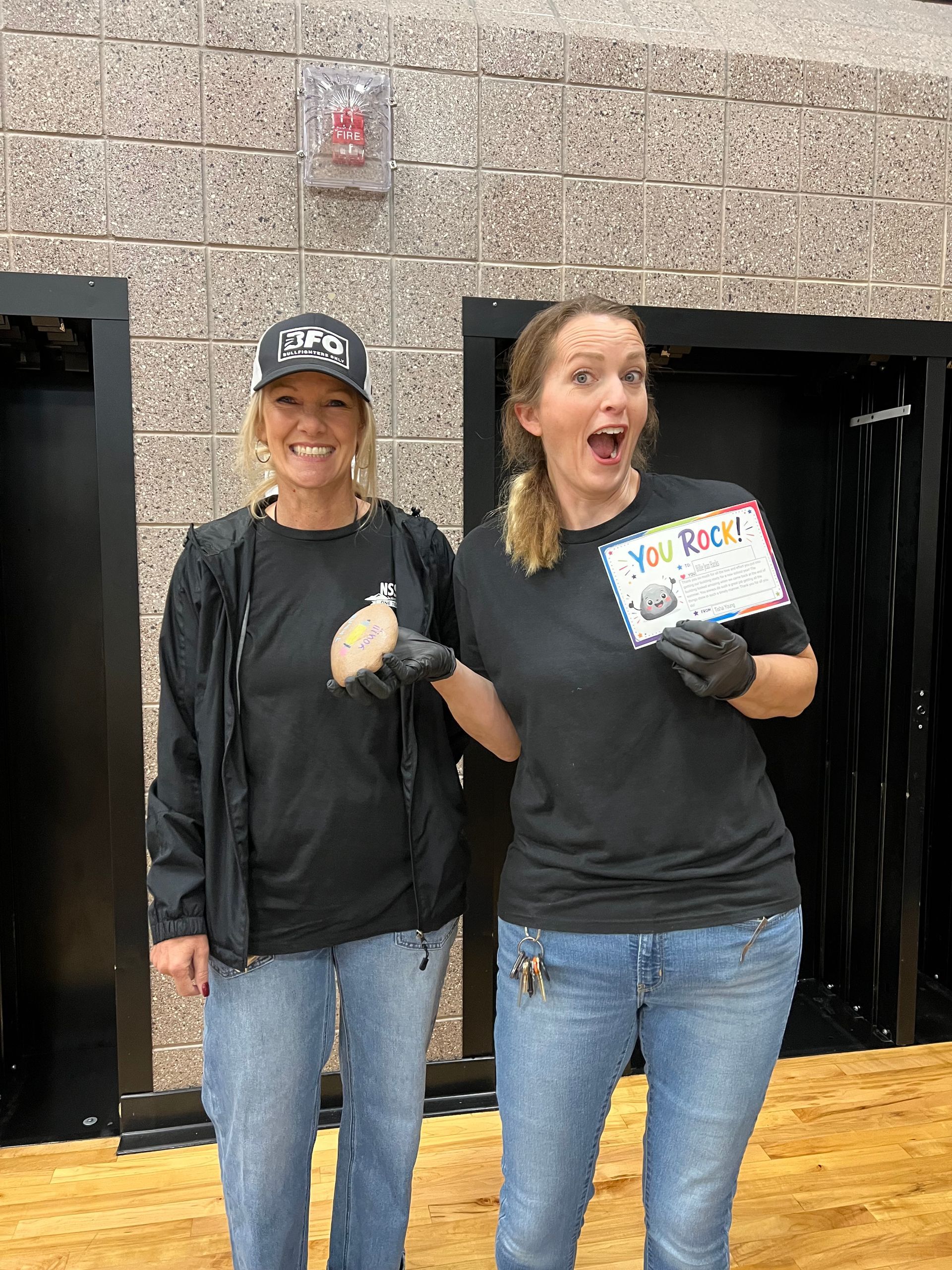 Two smiling people in black shirts and jeans stand in a doorway, one holding a colorful sign.