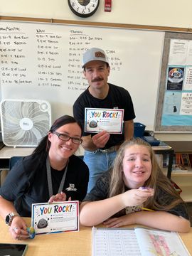 Three people in a classroom hold colorful “You Rock!” cards, smiling near a whiteboard.