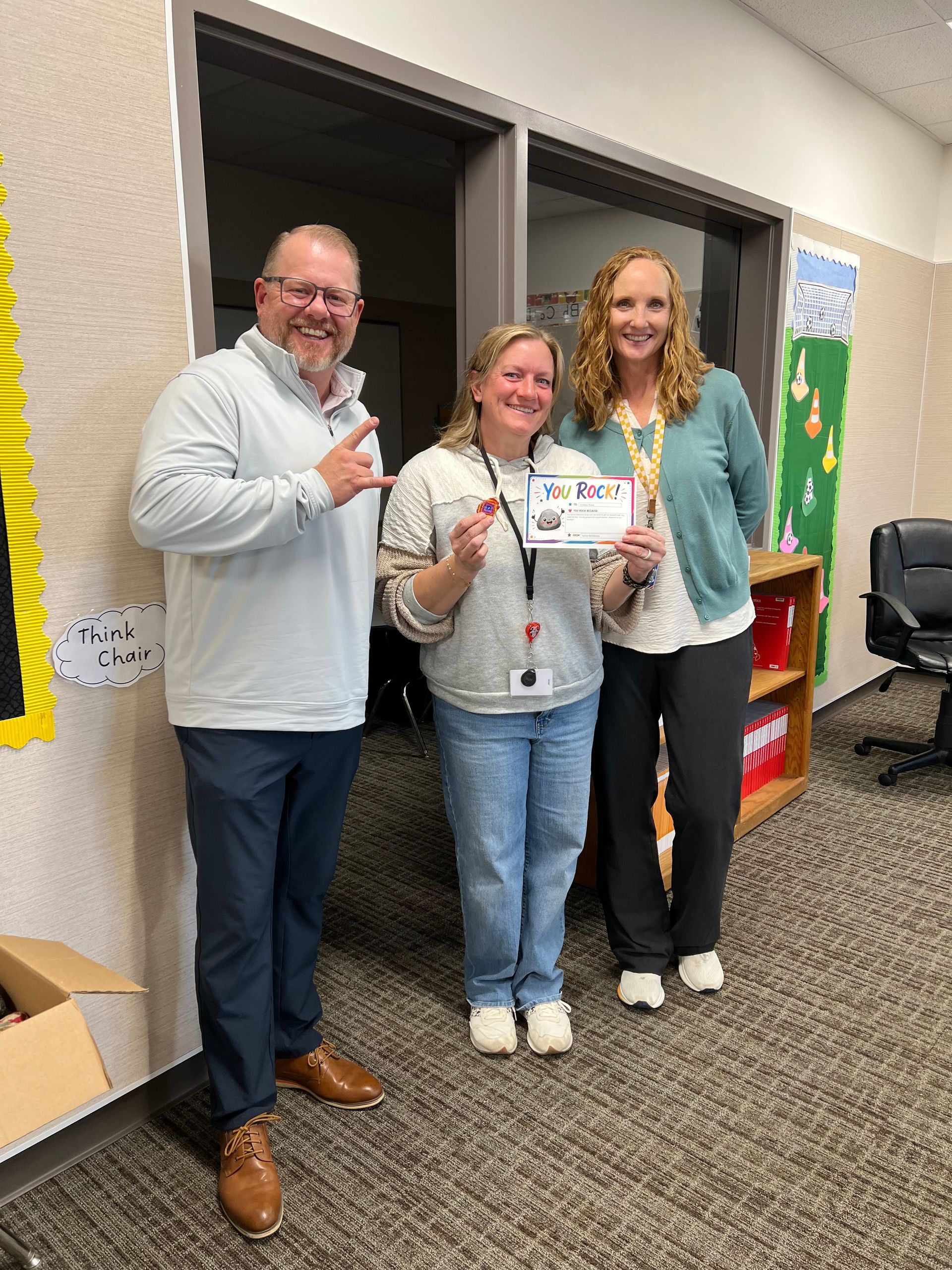 Three people smiling in a classroom hallway, holding a certificate or award.