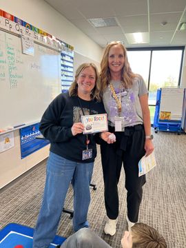 Two women smiling and holding a certificate in a classroom hallway