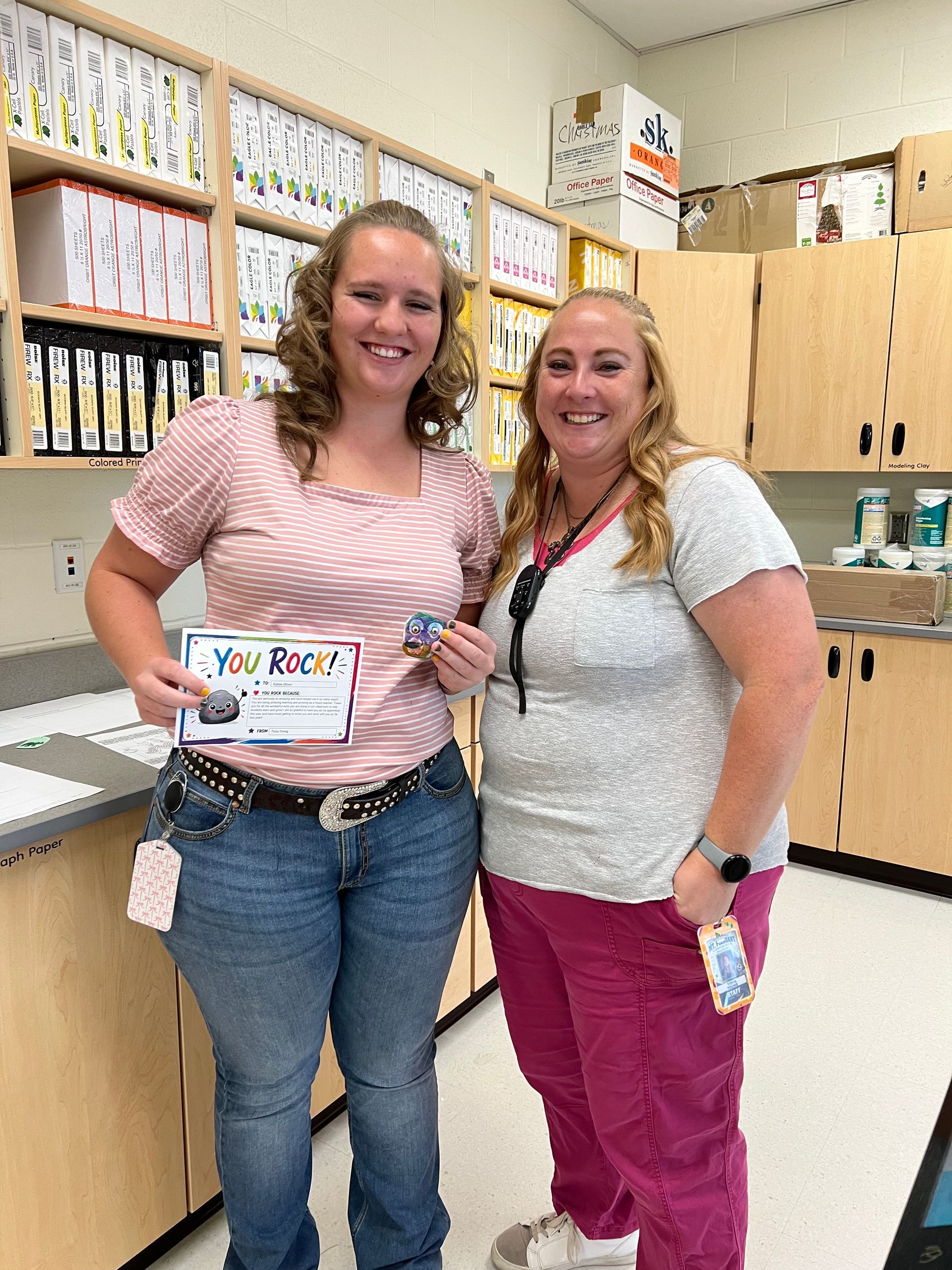 Two women smiling in a classroom, one holding a “You Rock!” card, with cubbies and papers behind them.