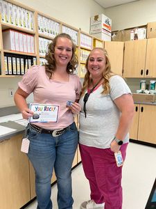 Two smiling coworkers stand in a store aisle, holding a certificate, with shelves of supplies behind them.