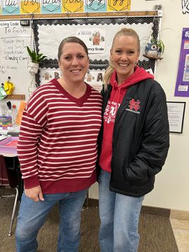 Two women smiling in a classroom, standing in front of a decorated bulletin board.