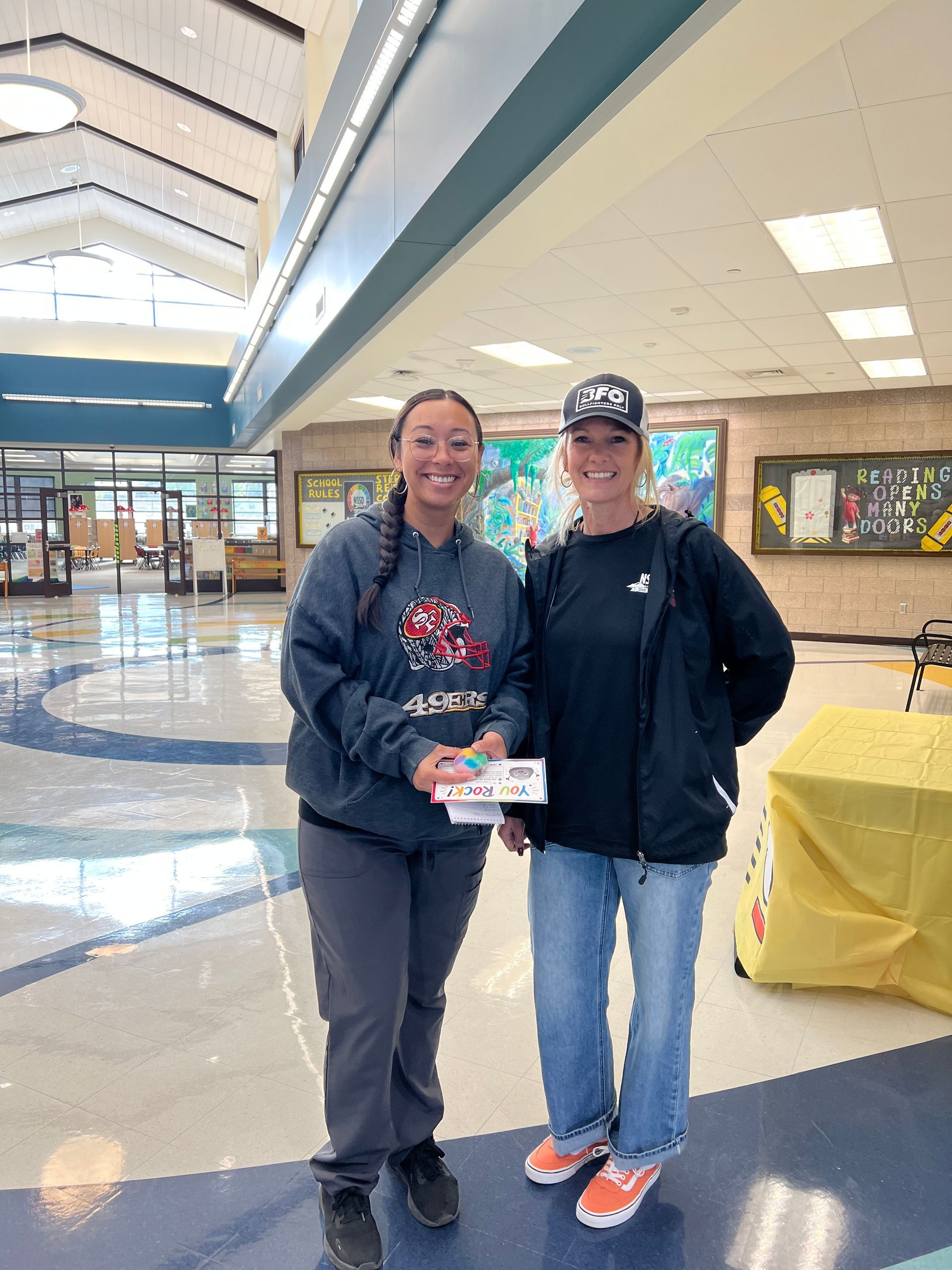 Two people posing in a school lobby, holding an item and smiling under a high ceiling.