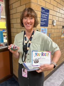 Smiling staff member in a hallway, holding a “You Rock!” box and a small decorated object.