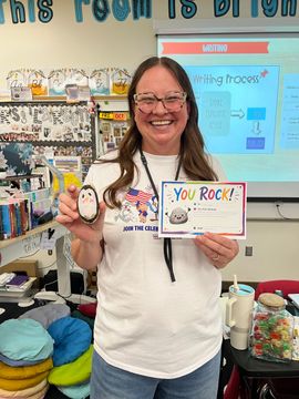 Smiling woman in a classroom holding a “You rock!” sign and a penguin craft display