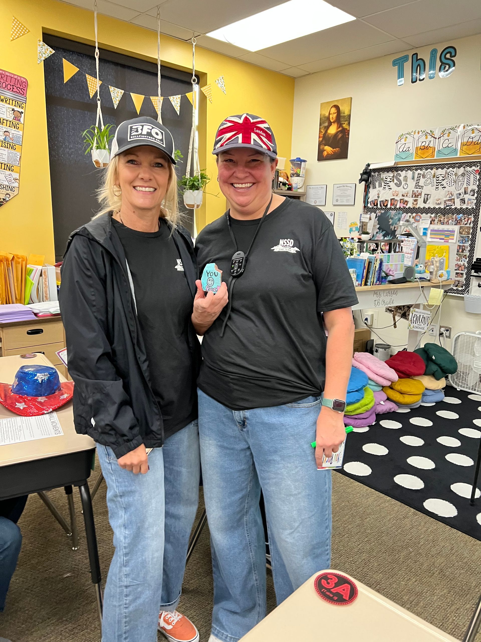 Two people posing in a craft store, wearing black tops and hats, with colorful supplies in the background