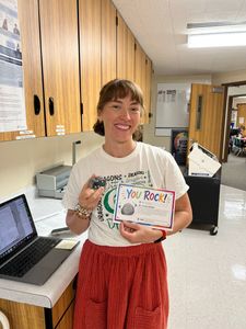 Smiling person in a room holding a “You Rock” card beside a laptop and wooden cabinets