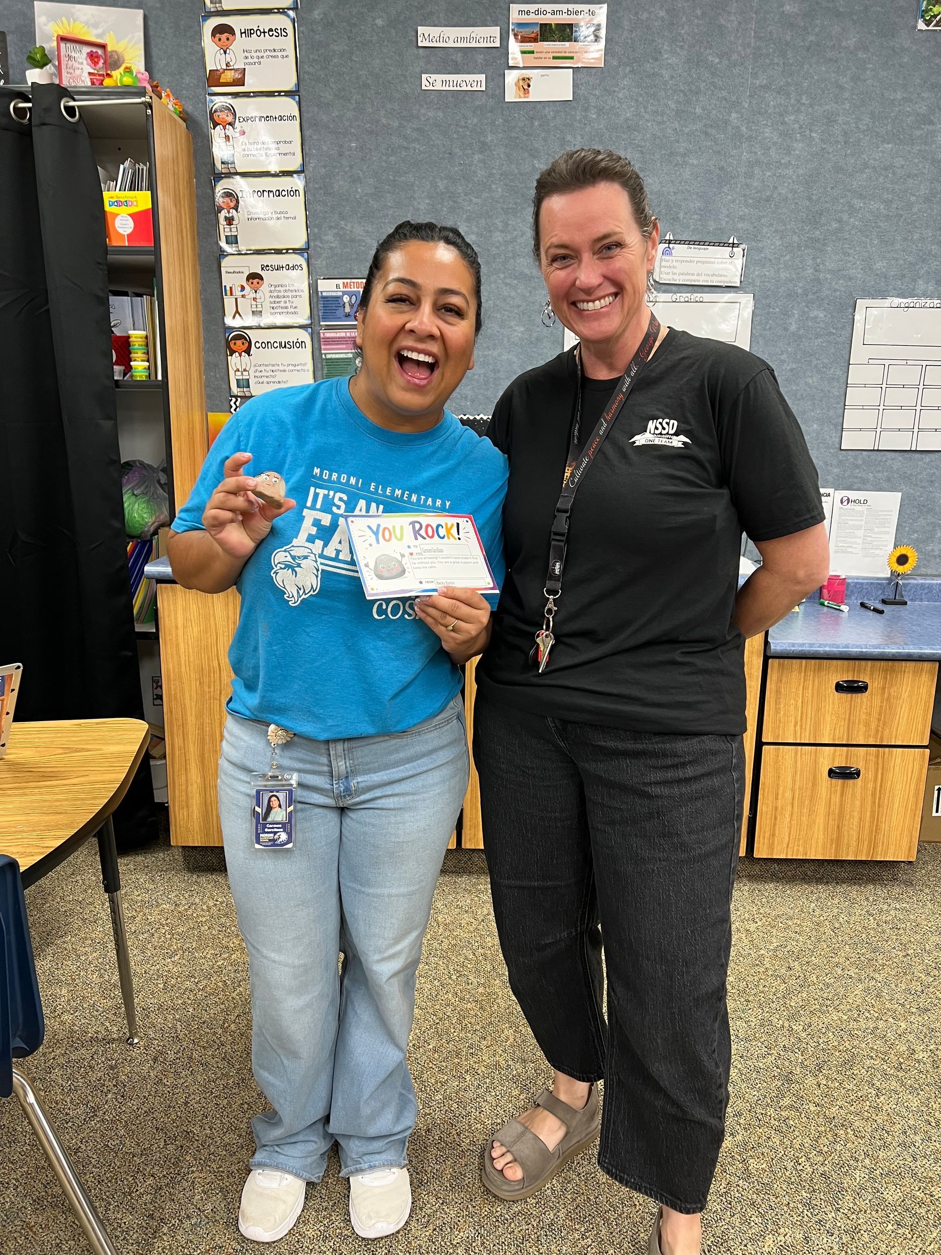 Two smiling people stand in a classroom, one holding a certificate, in front of a bulletin board.