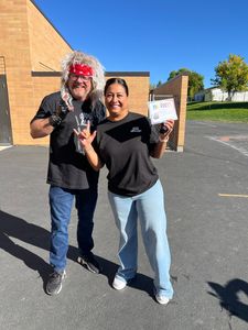 Two people smiling outdoors, holding papers and posing in a parking lot beside a brick building.