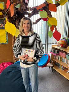 Woman holding a children’s book beside a colorful tree display in a classroom corner
