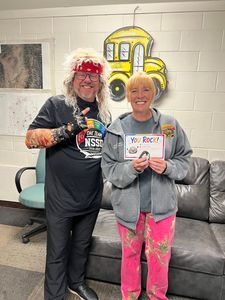 Two people smiling in a lounge, holding a sign, with a bright yellow wall decoration behind them