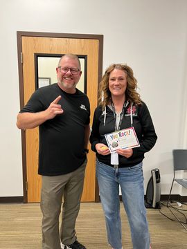 Two people posing indoors, one holding a certificate and both smiling near a doorway.