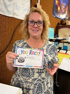 Woman holding a colorful “You Rock!” card in a craft room, smiling beside art supplies.