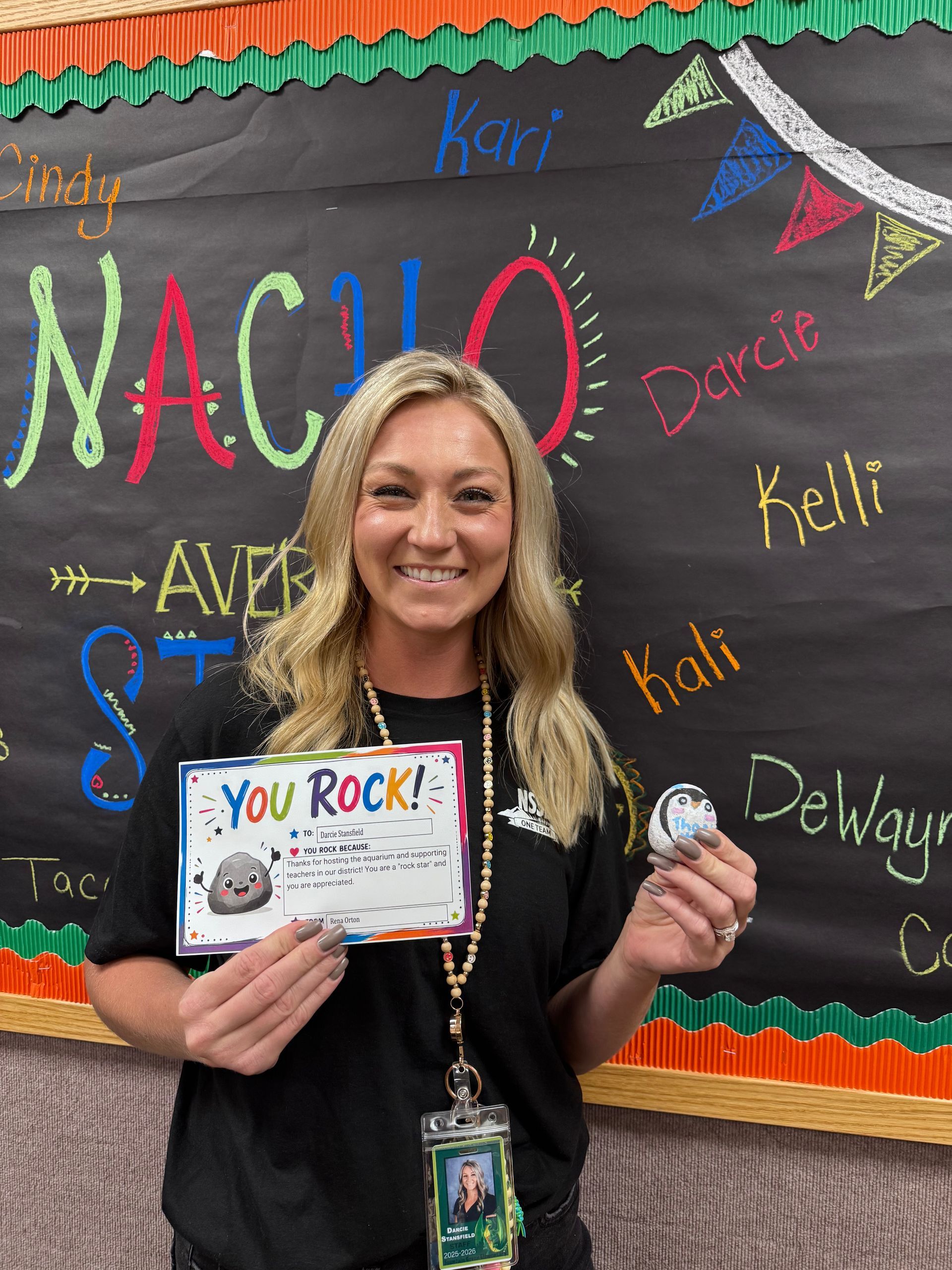 Smiling woman holds a “You Rock!” award in front of a colorful classroom chalkboard.