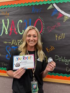 Teacher holding a “You Rock!” note in front of a colorful classroom bulletin board.