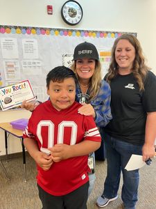 Three smiling people in a classroom, with a child in a red jersey in front and a “You Rock!” sign on the desk