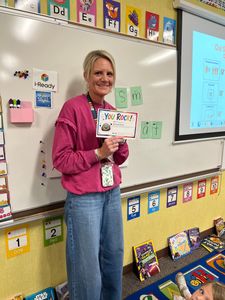 Teacher in pink sweater holding a book in a classroom beside a whiteboard and student work displays